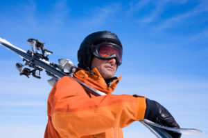 Man in an orange ski suit holding skis with bright blue sky behind him.