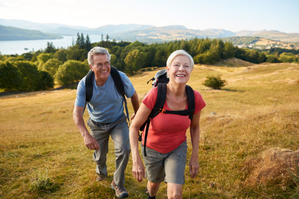 A middle-aged white couple wearing backpacks hikes up a hill overlooking a river.