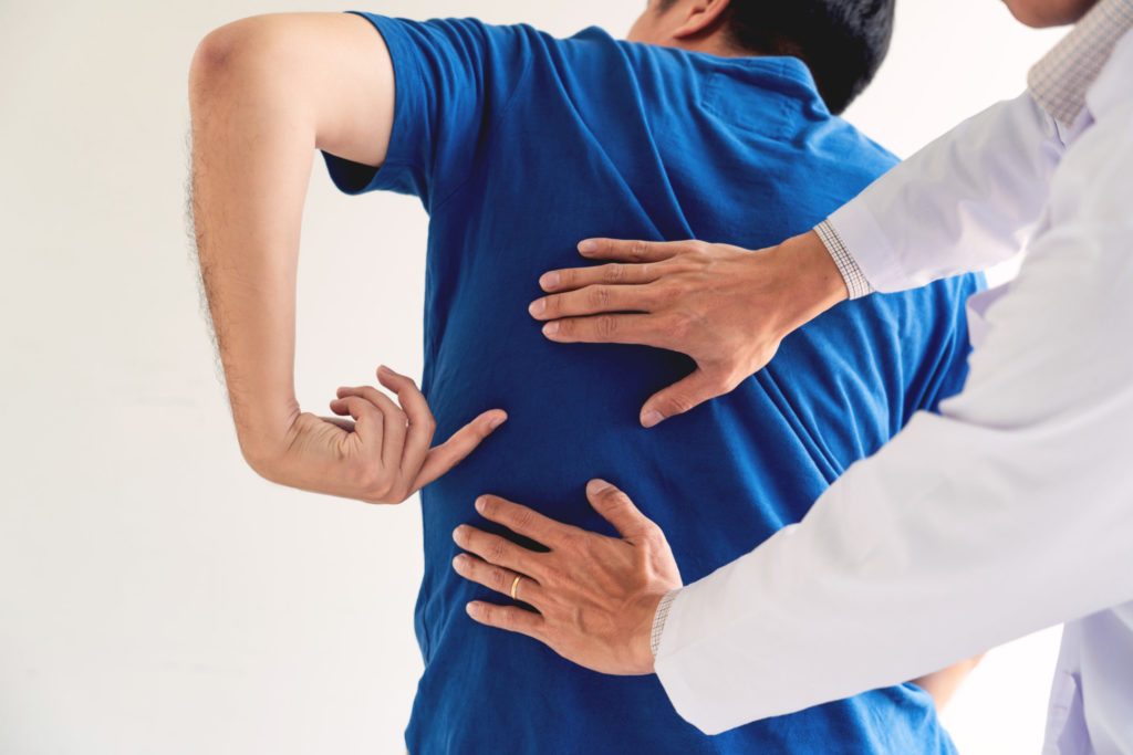 A man in a blue shirt points to a painful spot on his back while a doctor examines the area.