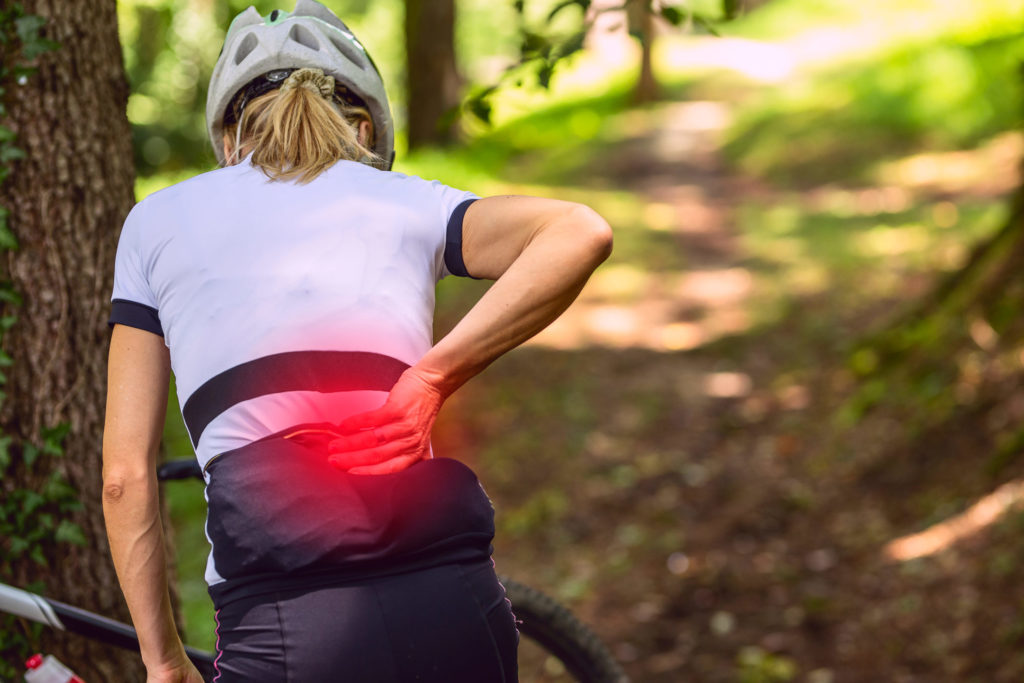 A female cyclist reaches for her lower back, which is highlighted in red to indicate pain.
