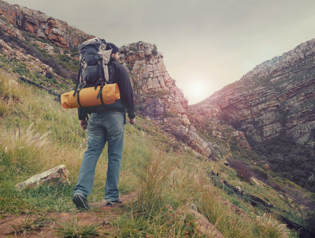 A man walks on a steep outdoor mountain path while wearing a heavy backpack. 