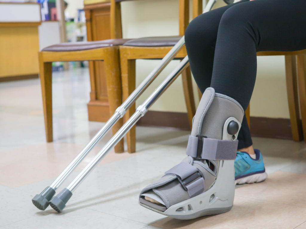 A patient sits in a waiting room with crutches and wearing a boot to protect her sprained ankle
