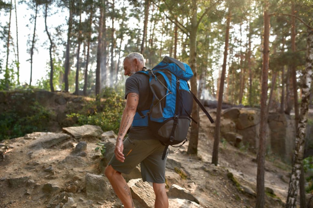 Older, fit white man hiking up a rocky hill in the forest with a large backpack on his back.