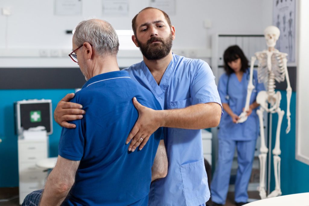 An orthopedic nurse helps an older male patient by stretching his shoulder and pressing on the center of his back.