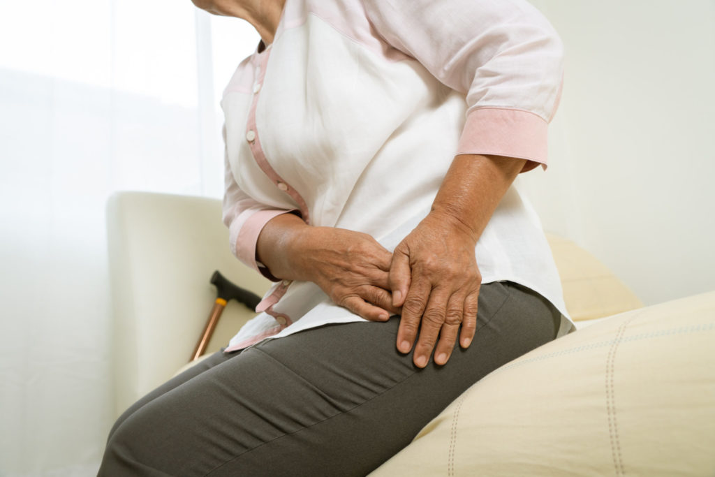 A middle-aged woman sitting on the edge of her couch with a cane next to her lays her hands on her painful hip.